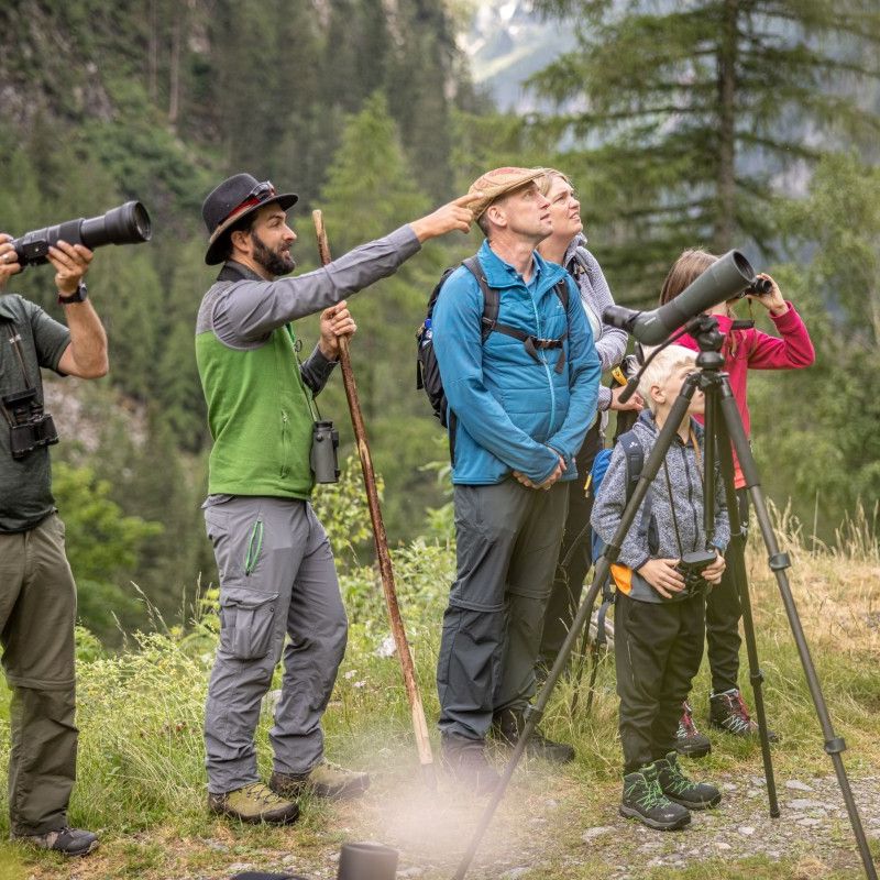 Ein Nationalparkranger mit einer Familie in der Natur. Er zeigt auf etwas. Daneben steht ein Mann mit einer Kamera